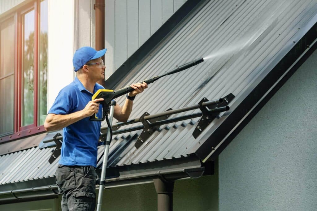 Service worker in a blue shirt and safety glasses pressure washing a corrugated metal roof with solar panel mounts