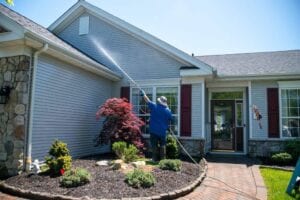 A professional cleaner in a blue shirt and hat, using a long wand to pressure wash the light gray vinyl siding and stone facade of a modern suburban home on a sunny day.
