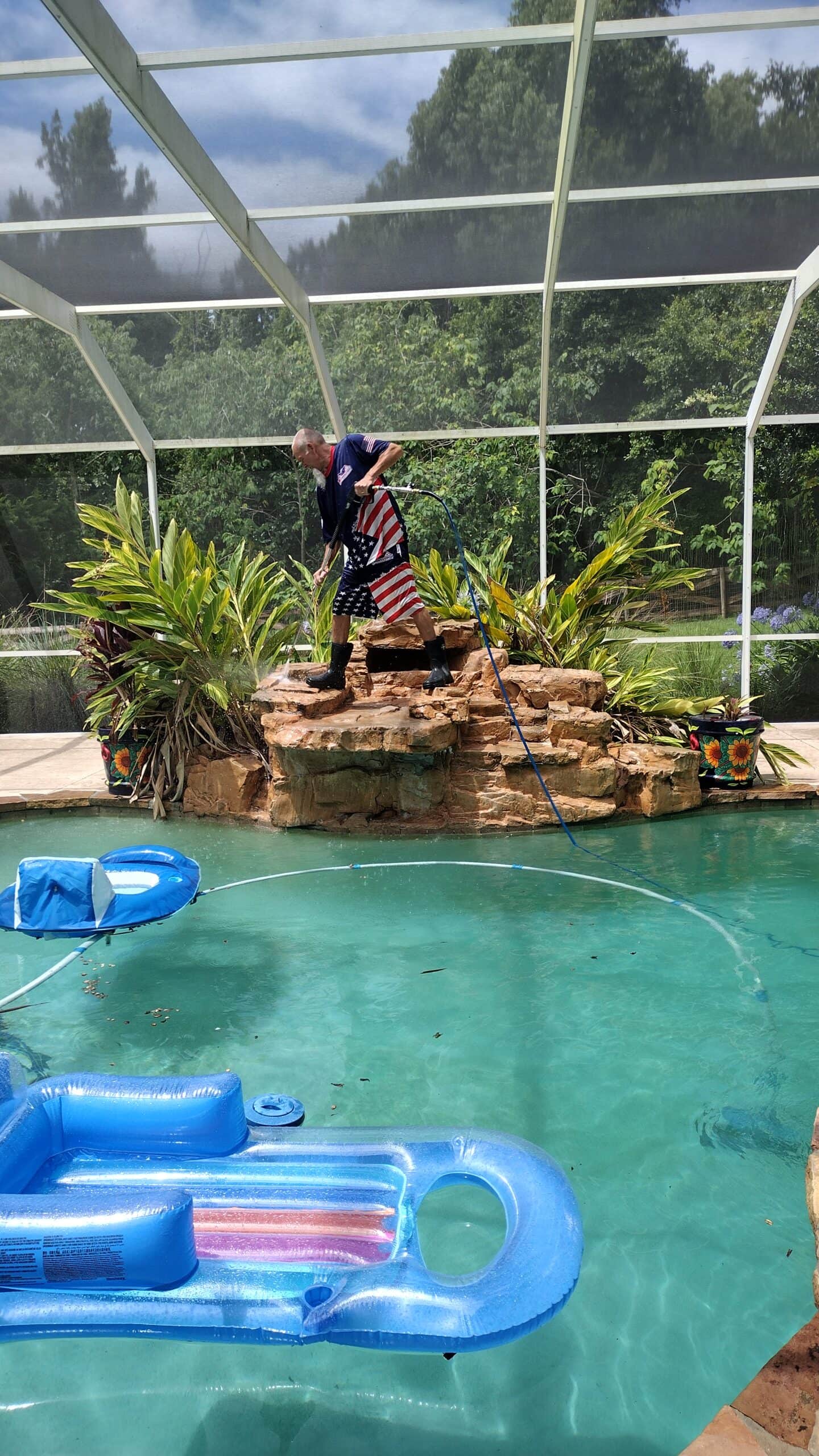 A man wearing shorts with an American flag pattern, boots, and a shirt is standing on a faux rock waterfall