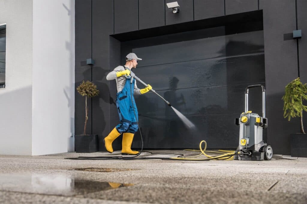 A professional cleaner in blue overalls, yellow boots and gloves, and a gray cap using a pressure washer to clean a large, glossy black garage door on a modern home, with the pressure washing machine visible on the concrete.