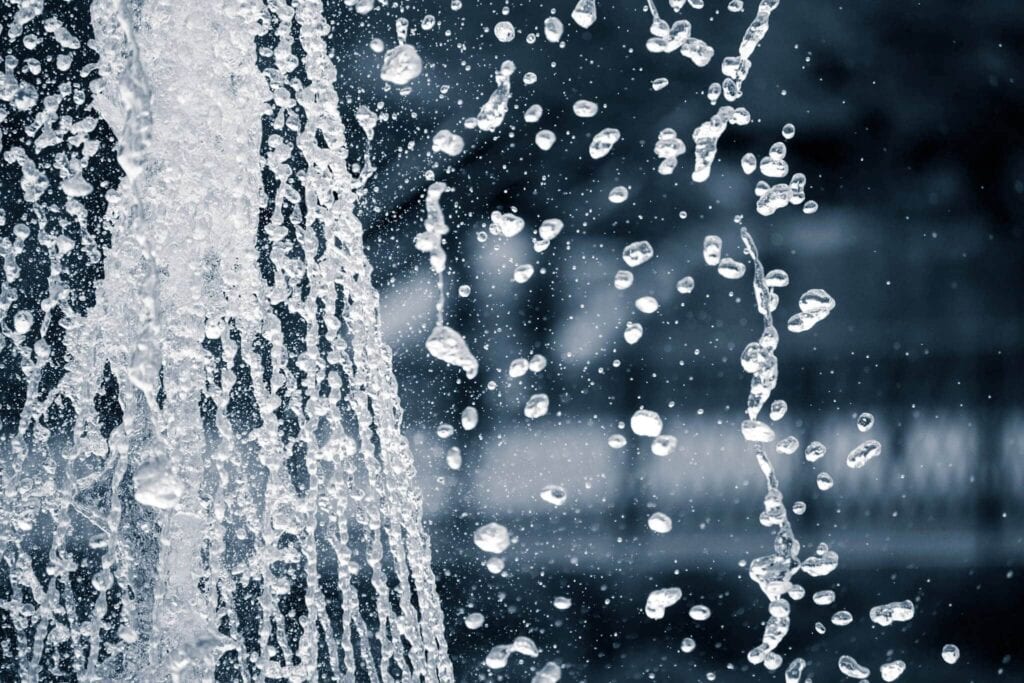 Close-up of clear water splashing and arcing in mid-air against a dark background, showing many individual water droplets in sharp focus.