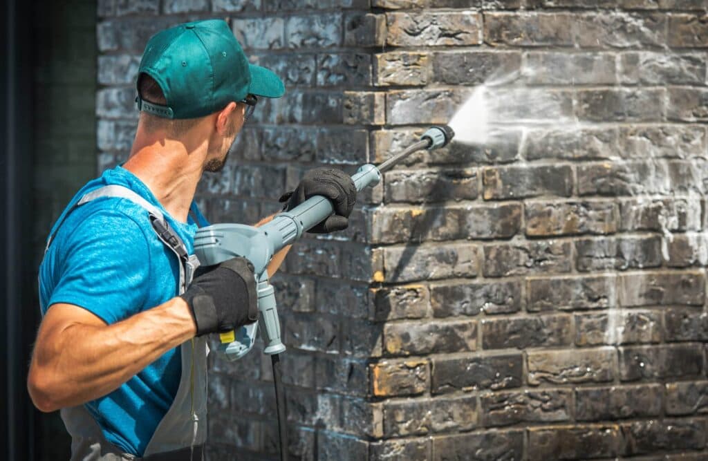 Close-up of a man in a blue shirt and green baseball cap power washing a dark gray or black brick column
