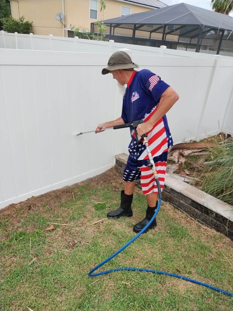 Older man in patriotic clothing and boots pressure washing a white vinyl privacy fence in a grassy yard.