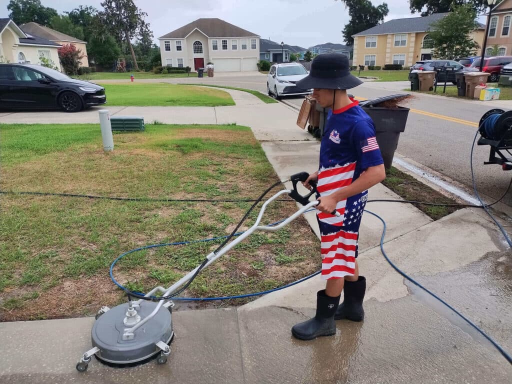 Pool cleaning technician in a hat and American flag shirt operating a rotary surface cleaner to pressure wash a wet concrete or paver driveway in a suburban neighborhood.