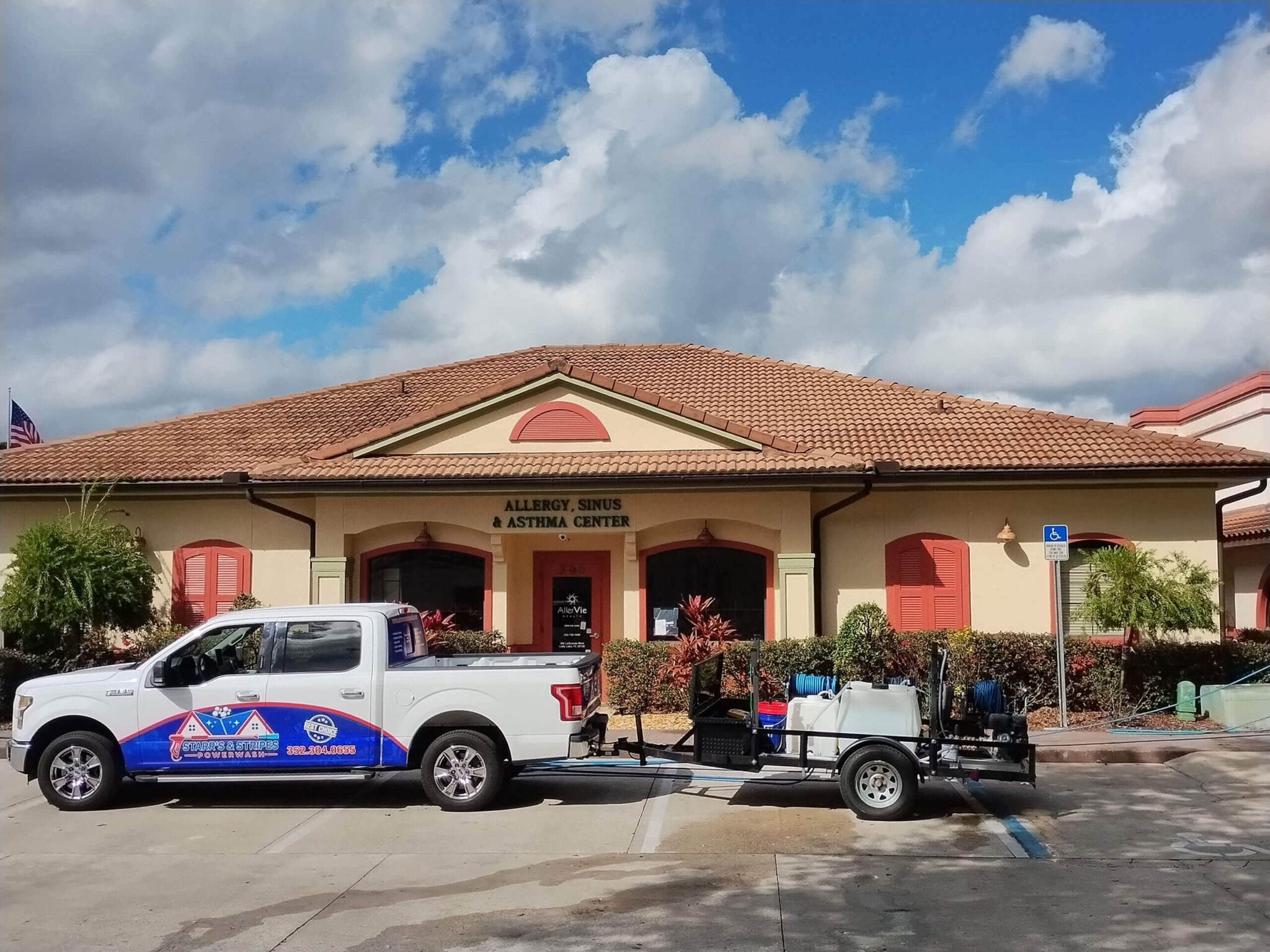 White pickup truck branded with a pool and home cleaning and maintenance company logo, towing a trailer with pressure washing and chemical tanks, parked outside a commercial building.