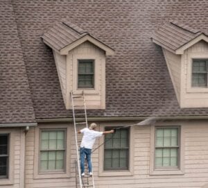 Worker standing on a ladder and using a pressure washer to clean the siding and trim around the dormer windows on a two-story house.