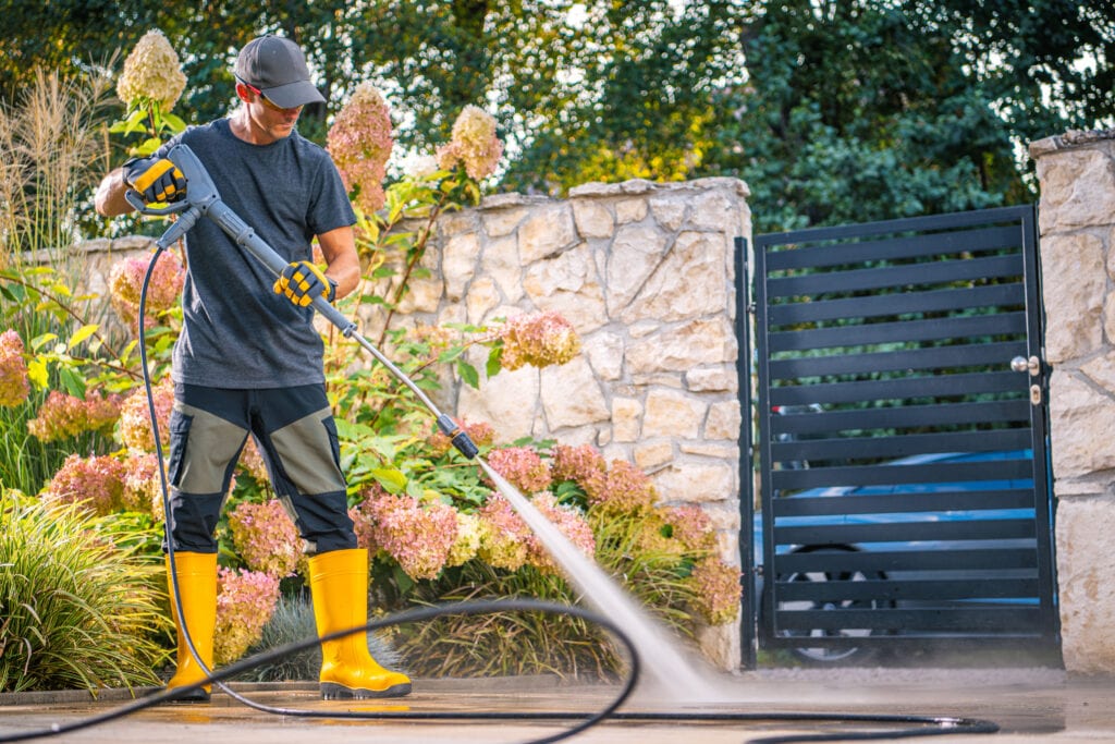 A professional worker in yellow safety boots and protective gear using a high-pressure washer to clean a stone driveway near a garden.