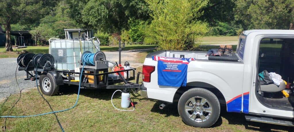 A white truck with a trailer carrying large water tanks and hoses on a grassy area. A blue and red shirt hangs on the truck. Background shows greenery.