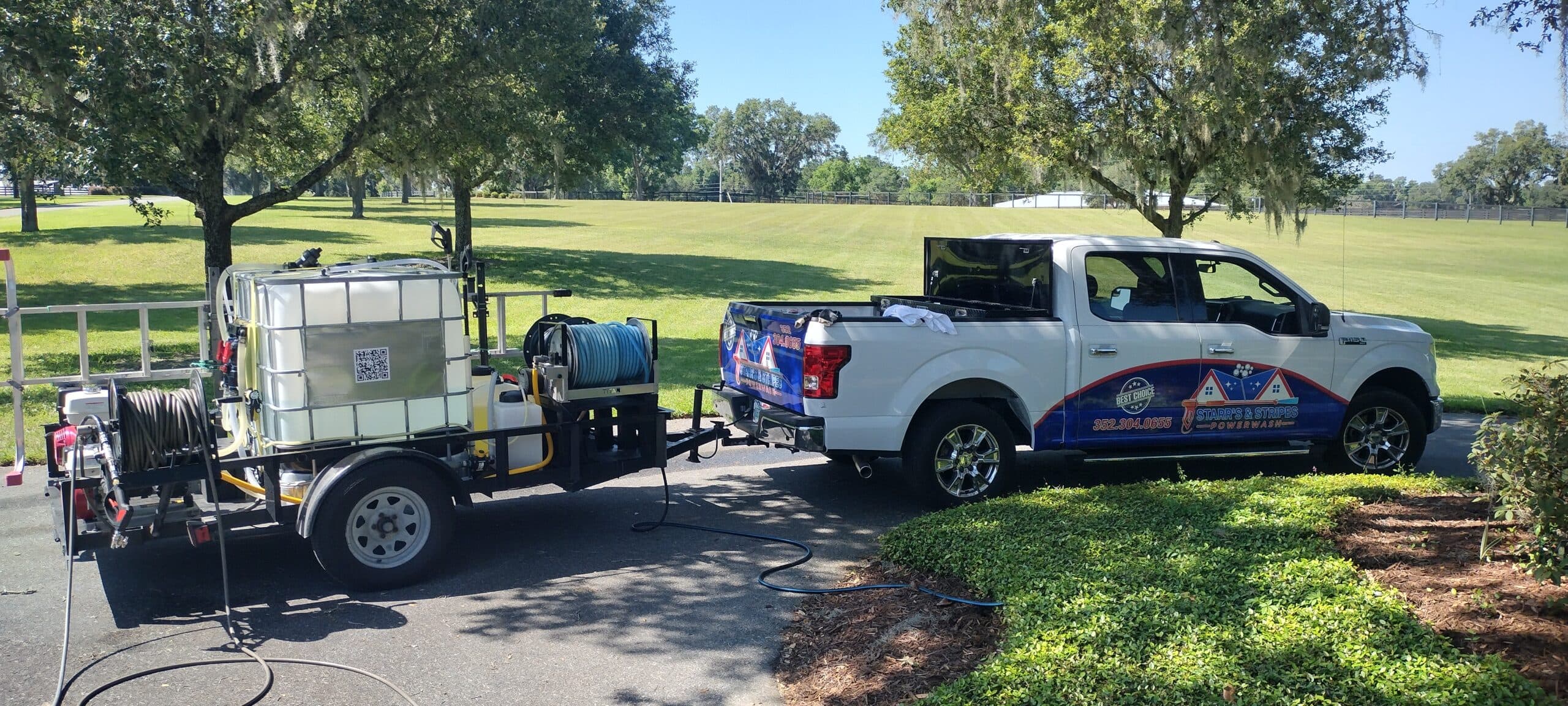A white pickup truck with a colorful logo pulls a trailer equipped with a large water tank and hoses. It's parked on a sunny street near green grass and trees.