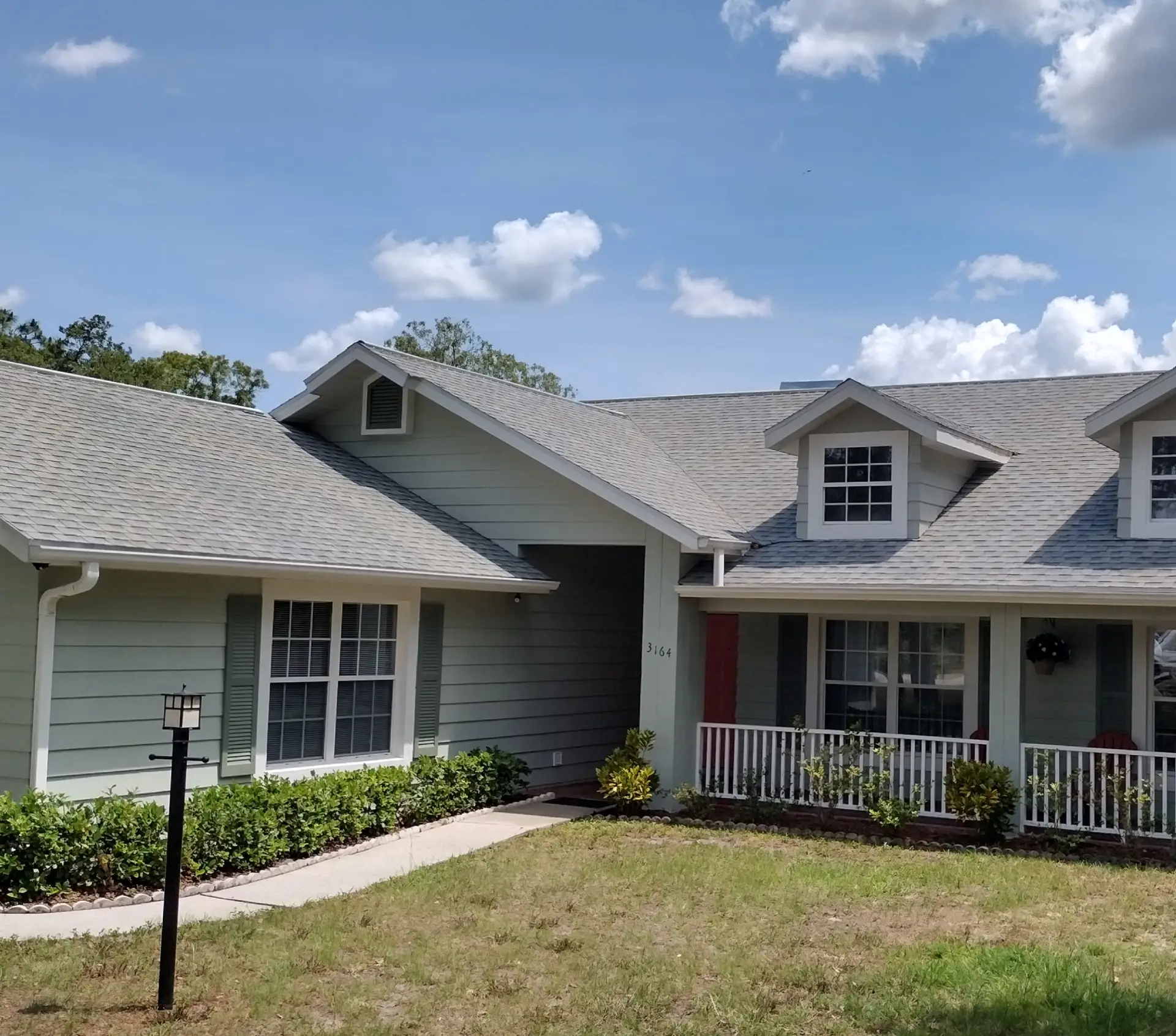 Single-story house with light gray siding and a gabled roof. Features include shuttered windows, a small porch, lush hedge, and a blue sky overhead.