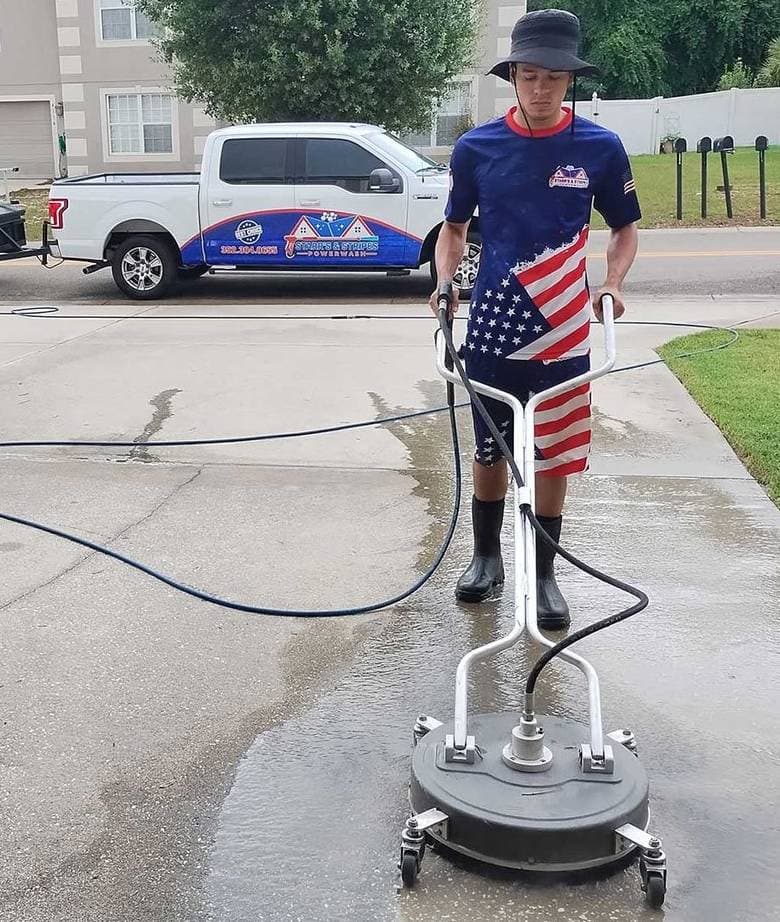 A man wearing American flag-themed shorts and a navy shirt operates a surface cleaner on a concrete driveway. A branded service truck and trailer are in the background, with suburban houses and green lawns around, under a clear sky.