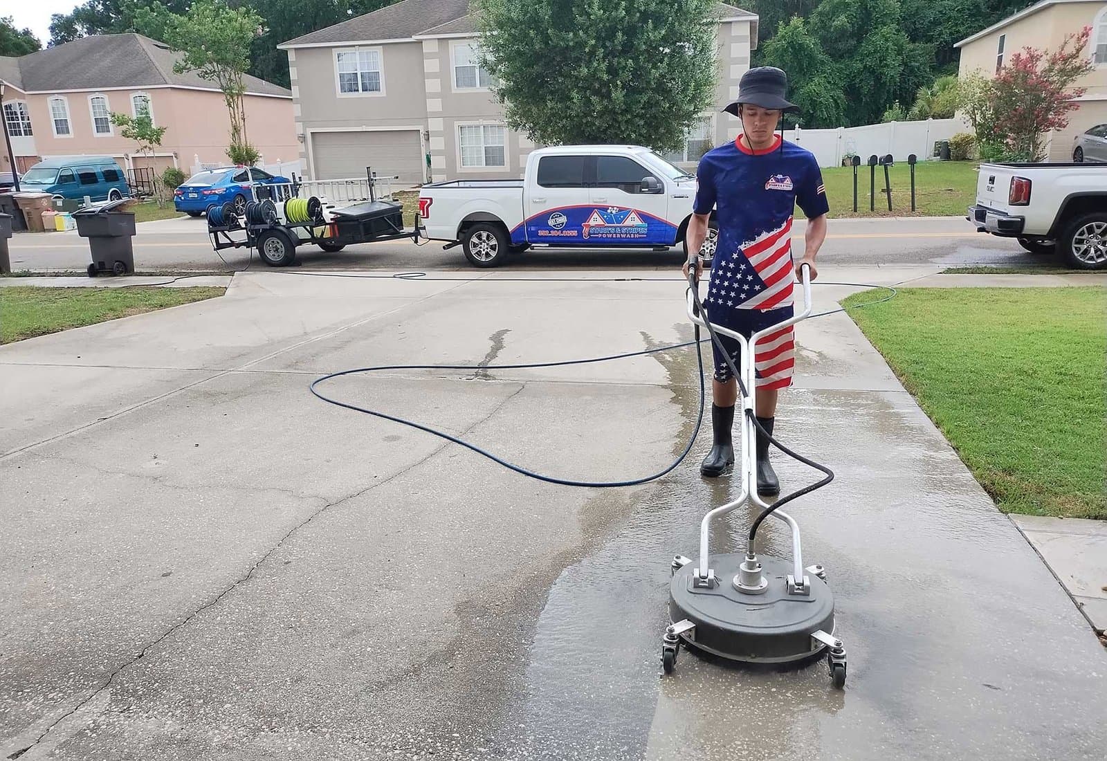 A man wearing American flag-themed shorts and a navy shirt operates a surface cleaner on a concrete driveway. A branded service truck and trailer are in the background, with suburban houses and green lawns around, under a clear sky.