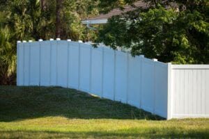 A long, clean white vinyl privacy fence installed on a grassy slope surrounding a residential home with lush green trees in the background.