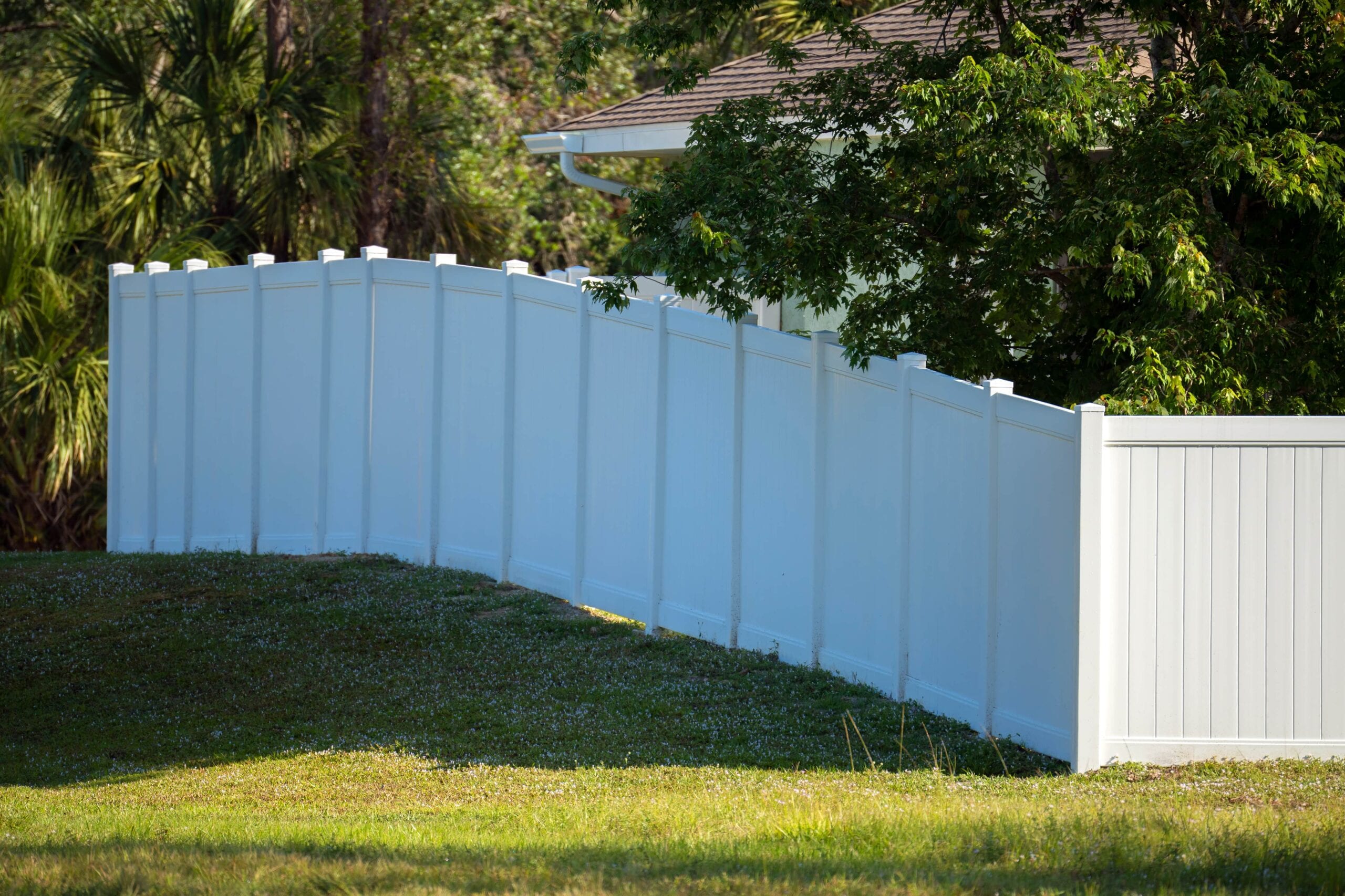 A long, clean white vinyl privacy fence installed on a grassy slope surrounding a residential home with lush green trees in the background.
