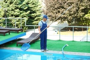 A male pool technician in blue overalls and a cap using a long-handled blue scoop net to clean debris from a clear outdoor swimming pool at a resort or residential property.