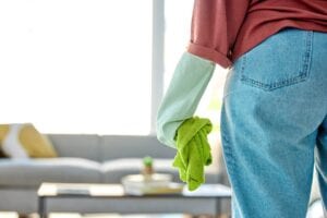 Rear view of a person wearing a light blue protective glove and holding a bright green microfiber cleaning cloth in a modern, sunlit living room.