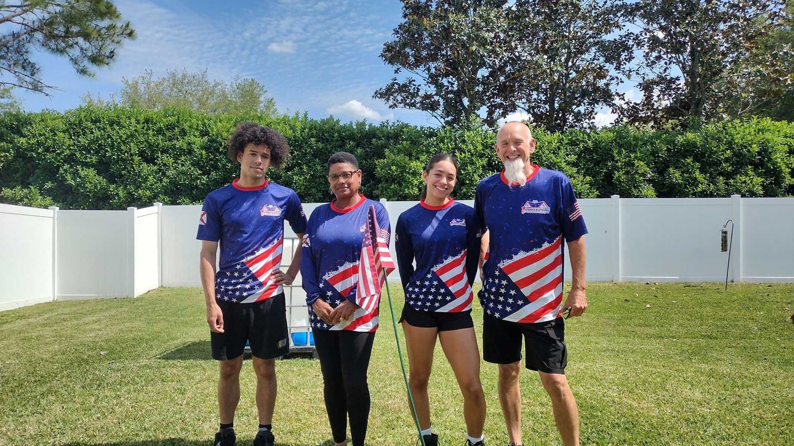 Four people standing on grass in a backyard, wearing matching blue shirts with American flag patterns, posing for a group photo.