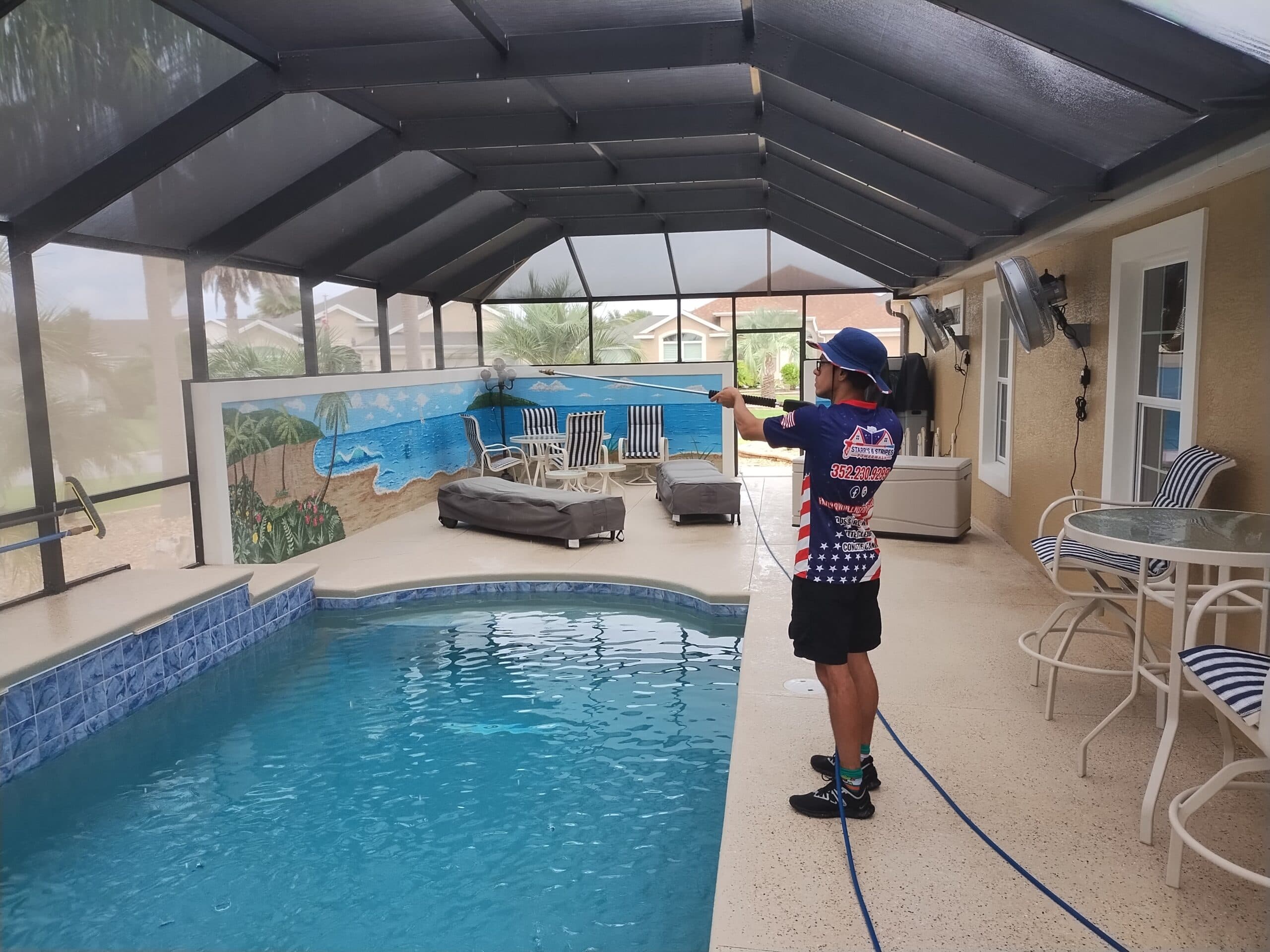 A person in a blue hat and patterned shirt cleans a covered pool area with a pressure washer. The area includes a mural, lounge chairs, and a glass table.