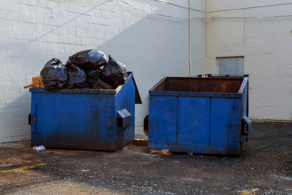 Two blue commercial metal dumpsters positioned against a white brick wall in a back alley; one bin is overflowing with black plastic garbage bags.