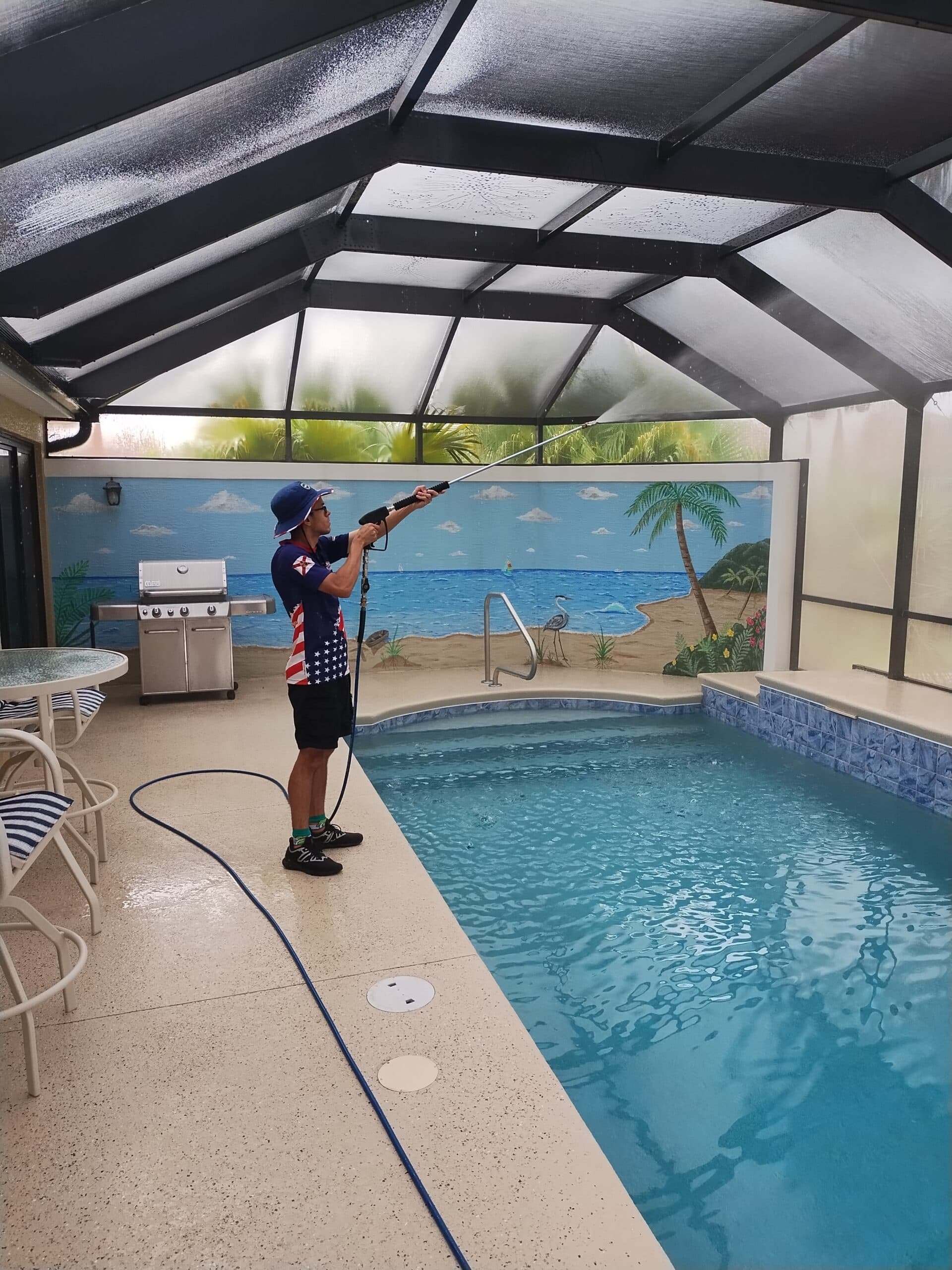 A person pressure washes the ceiling of an enclosed pool area. The scene is bright, with a mural of a beach and palm trees in the background.