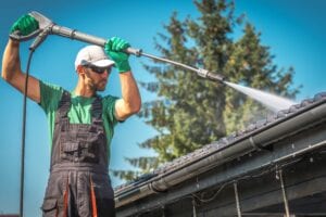 A professional cleaner in overalls and safety gear using a high-pressure wand to wash a corrugated plastic carport roof and gutters against a blue sky.