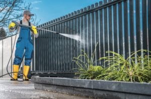 A professional cleaner in blue overalls, yellow boots, and safety gear using a high-pressure washer to clean a large black metal residential gate and fence.
