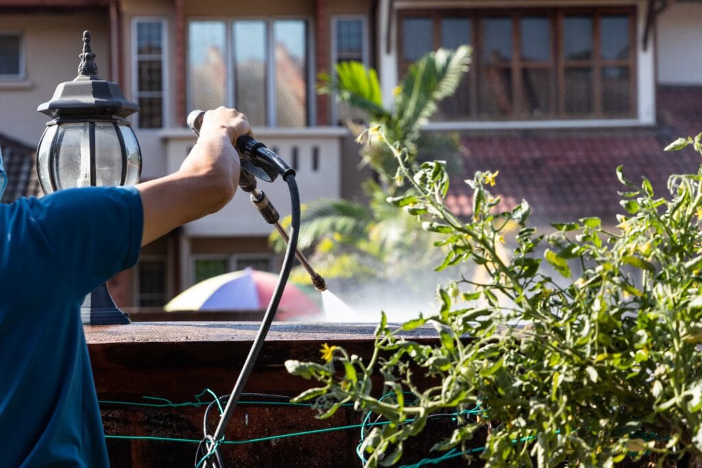 A professional worker in a blue shirt using a high-pressure water jet spray gun to clean a residential roof surface near garden plants.