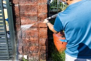 A close-up shot of a person using a high-pressure water jet spray gun to clean dirt and grime off a textured red brick gate pillar.
