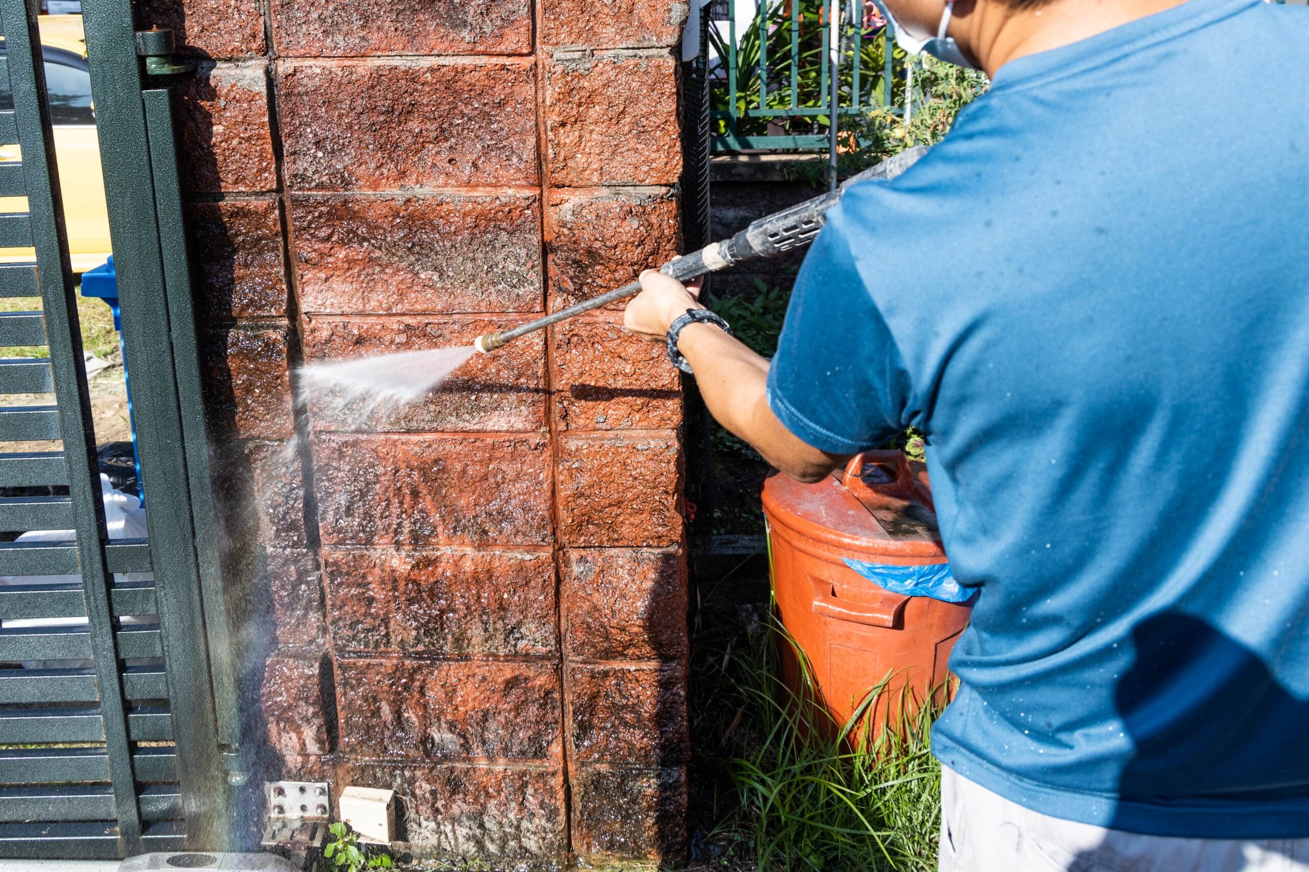 A close-up shot of a person using a high-pressure water jet spray gun to clean dirt and grime off a textured red brick gate pillar.