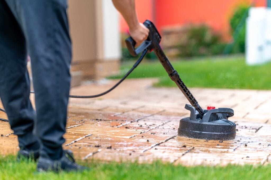 Close-up of a person using a circular pressure washer surface cleaner attachment to remove dirt and grime from a stone paver patio.