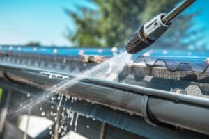 A close-up action shot of a pressure washer nozzle spraying a powerful stream of water and foam into a black metal rain gutter to remove debris.