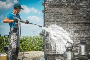 A professional technician in grey protective gear uses a high-pressure washer to remove white stains and buildup from a dark brick exterior wall during a residential cleaning service in Orlando, FL.