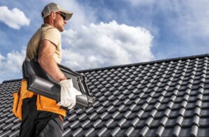 A professional roofer carrying heavy black ceramic tiles during a new roof installation service in Austin, TX, under a blue sky.